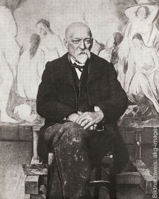 Fotografía histórica en blanco y negro del pintor francés Paul Cézanne, sentado en su estudio frente a un gran cuadro con representaciones de desnudos.