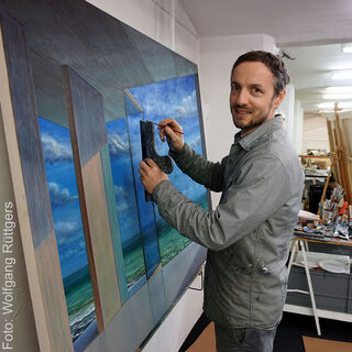 Der Maler Tobias Stutz blickt lächelnd in die Kamera, während er im Atelier an einem Bild arbeitet.
