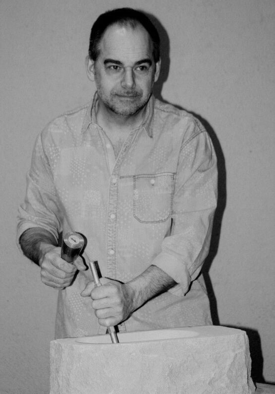 Fotografia in bianco e nero dello scultore belga Marc Caelenberghe mentre lavora con scalpello e martello su una scultura in pietra.