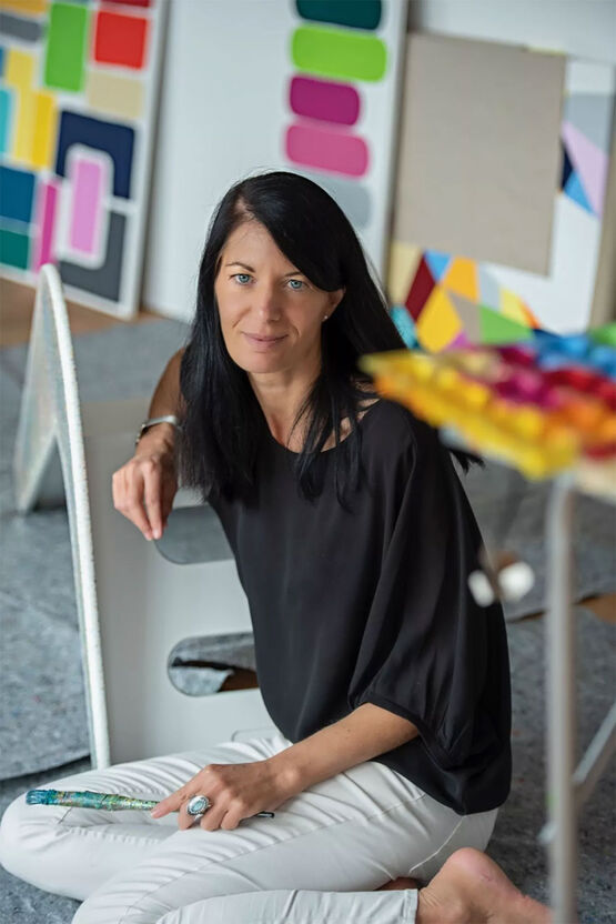 La peintre Astrid Stoeppel est assise par terre dans son atelier, devant des tableaux colorés, un pinceau à la main.