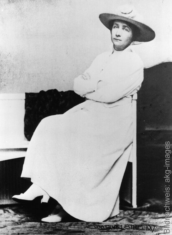 Historische Schwarz-Weiß-Fotografie der Malerin Gabriele Münter in hellem Kleid und Hut mit verschränkten Armen sitzend.