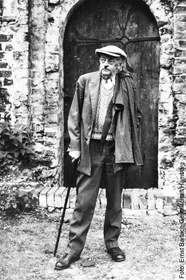 Le sculpteur allemand Ernst Barlach se tient devant une vieille porte en bois, muni d'une canne et coiffé d'une casquette.