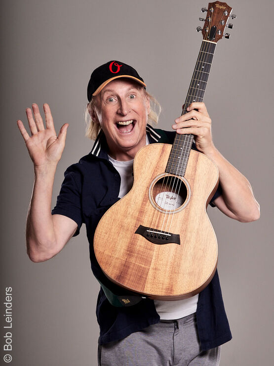 Otto Waalkes, le Frison oriental le plus célèbre et artiste, pose joyeusement avec une guitare acoustique et fait un signe de la main à la caméra.