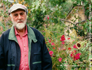 Der österreichische Künstler und Maler Friedensreich Hundertwasser mit Mütze und Bart stehend in einem blühenden Garten mit rosa Blumen.