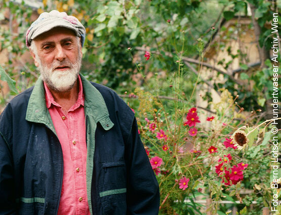 Der österreichische Künstler und Maler Friedensreich Hundertwasser mit Mütze und Bart stehend in einem blühenden Garten mit rosa Blumen.