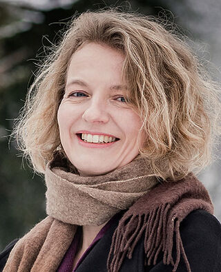 La pittrice Kathrin May, con i capelli biondi e ricci e una sciarpa marrone, guarda sorridente verso la fotocamera.