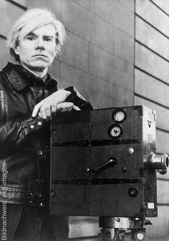A black-and-white photograph of the Pop Art artist Andy Warhol, wearing a leather jacket, standing next to a vintage film camera.