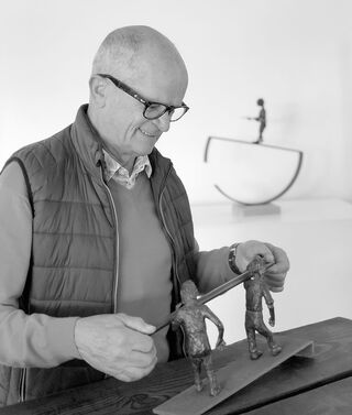 Lo scultore belga Freddy De Waele osserva, sorridendo, le sue delicate sculture in bronzo posate su un tavolo di legno.