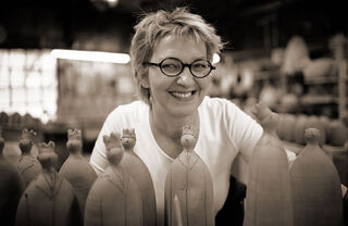 Portretfoto van de kunstenares Susanne Boerner, die vrolijk glimlachend tussen haar handgemaakte kleisculpturen in haar atelier poseert.