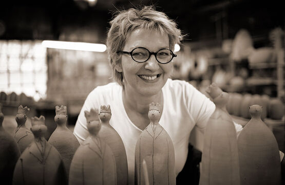 Retrato de la artista Susanne Boerner, que posa con una sonrisa alegre en medio de sus esculturas de arcilla hechas a mano en su taller.
