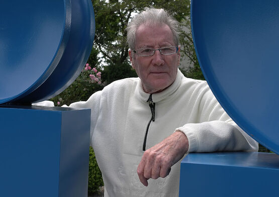 L'artista Renaat Ramon, con gli occhiali e un maglione di pile bianco, guarda attraverso l'apertura di una grande scultura blu.