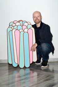 Daniel Engelberg è inginocchiato, sorridente, accanto a un grande oggetto geometrico colorato appeso alla parete.