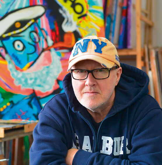 L'artiste Holger Mühlbauer-Gardemin, portant des lunettes et une casquette de baseball new-yorkaise, regarde droit devant lui devant une œuvre d'art colorée.