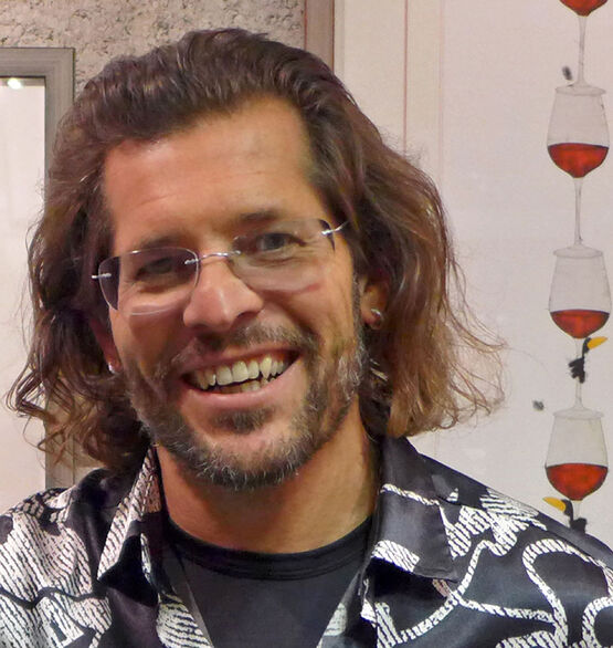 Porträt des österreichischen Künstlers, Malers und Zeichners Michael Ferner lächelnd mit Brille vor einem seiner Kunstwerke mit Weingläsern.