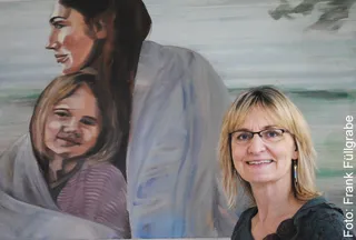 Kunstneren og maleren Anja Struck står smilende foran et af sine oliemalerier, der forestiller to personer.