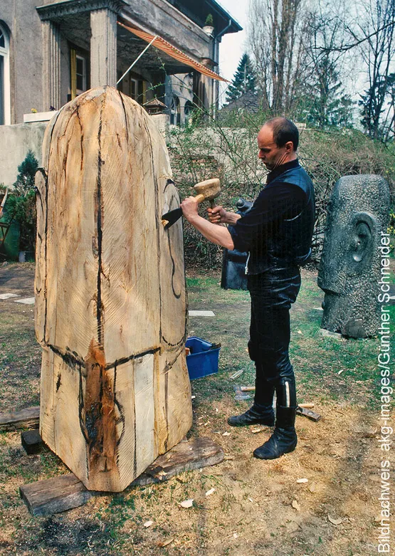 Lo scultore Rainer Kriester sta lavorando all’aperto a una grande scultura in legno, utilizzando un martello da intaglio e uno scalpello.