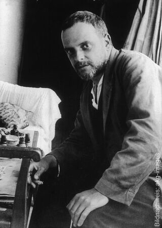 A historical black-and-white photograph of the German-Swiss artist and painter Paul Klee, bearded, sitting in his studio next to a table.