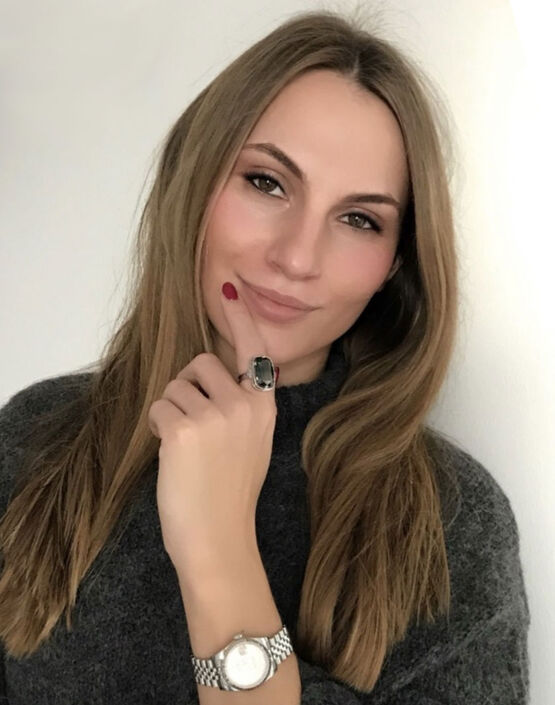 Portrét ruské umělkyně a malířky Olgy Jonasové, která se usmívá v šedém svetru s rukou na bradě před světlým pozadím.