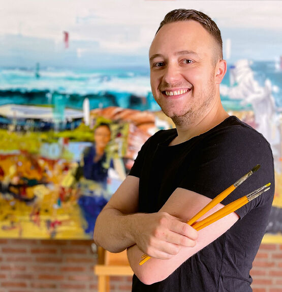 Současný malíř Michael Bajer stojí s úsměvem, se založenýma rukama a štětci v rukou, před velkým uměleckým dílem.