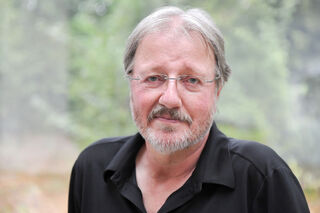 Un portrait de l'artiste allemand Heiner Thiel, portant des lunettes et une barbe, sur un fond clair.