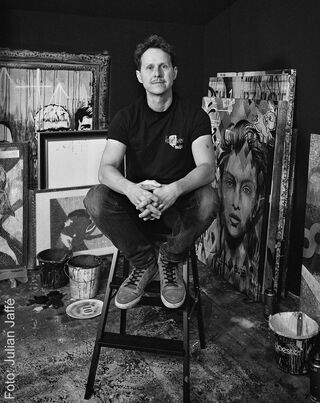 Ritratto in bianco e nero dell’artista BUJA seduto su una scaletta nel suo studio, circondato da quadri.