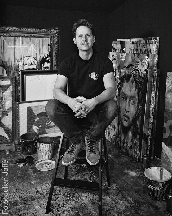 Zwart-witportret van de kunstenaar BUJA, zittend op een trapladder in zijn atelier, omringd door schilderijen.