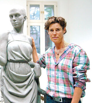 La scultrice Kristin Kolb, con indosso una camicia a quadri, sorride nel suo luminoso atelier accanto a una scultura a grandezza naturale di una donna in argilla grigia da lei realizzata.