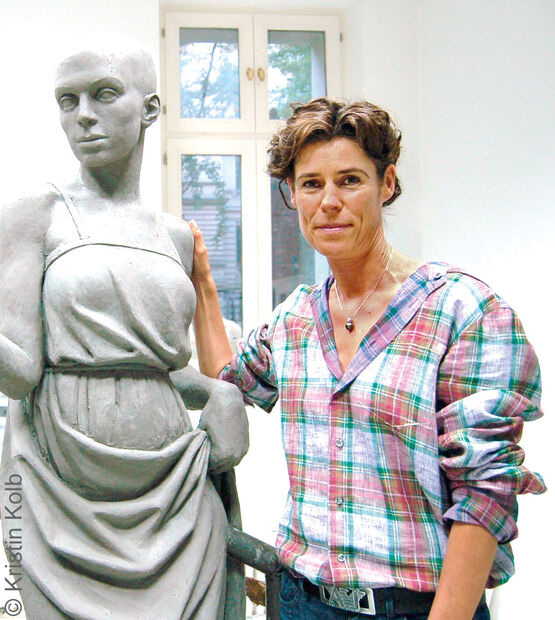 Dans son atelier lumineux, la sculptrice Kristin Kolb, vêtue d'une chemise à carreaux, sourit à côté d'une sculpture en argile grise représentant une femme en taille réelle qu'elle a réalisée.