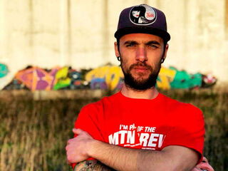 A portrait of the graffiti artist Bruno Jorge Monteiro e Silva, wearing a red T-shirt and a cap, standing in front of a graffiti-covered wall.