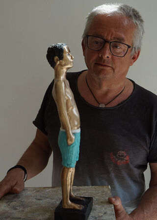 Den tyske kunstner og billedhugger Peter Hermann, der bærer briller, betragter koncentreret sin skulptur af en dreng i badebukser.