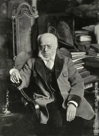 Historische Schwarz-Weiß-Fotografie des Historienmalers Adolph von Menzel mit Brille sitzend in einem Sessel.