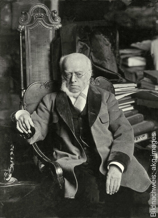 Historische Schwarz-Weiß-Fotografie des Historienmalers Adolph von Menzel mit Brille sitzend in einem Sessel.