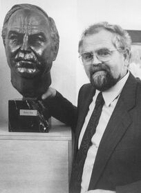 Der Bildhauer Kurt Arentz posiert im Anzug lächelnd neben seiner bronzenen Porträtbüste von Altbundeskanzler Helmut Kohl.