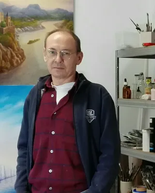 Der surrealistische Maler Viktor Lau steht mit Brille und dunkelblauer Jacke in seinem Atelier vor Ölgemälden.