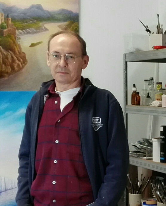 Surrealistický malíř Viktor Lau stojí ve svém ateliéru před olejomalbami, má na sobě brýle a tmavě modrou bundu.