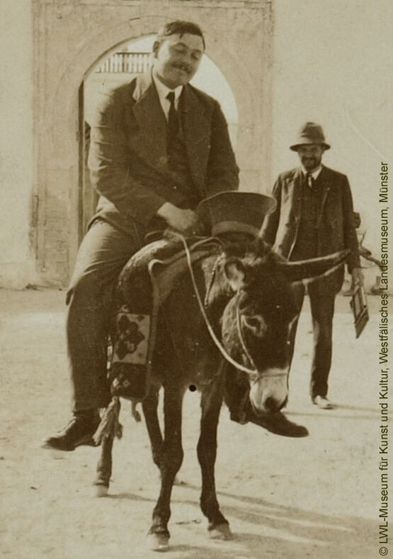 Historische Schwarz-Weiß-Fotografie des Malers August Macke im Anzug auf einem Esel reitend.