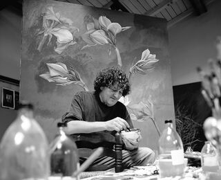 A black-and-white photograph of the Italian artist Bruno Bruni at work in his studio in front of a large painting featuring floral motifs.