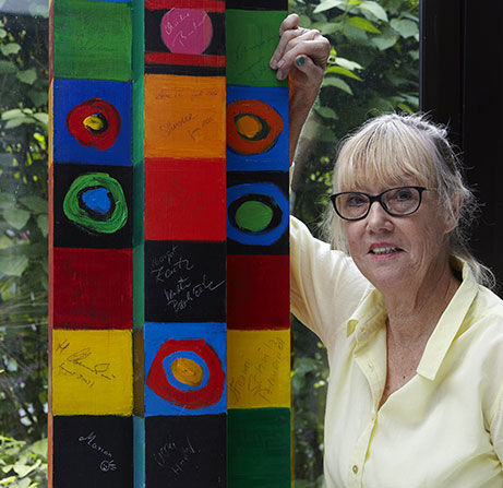 L'artista e pittrice Christine Kremkau, con gli occhiali, è in piedi accanto a una stele decorata con motivi colorati da lei stessa.