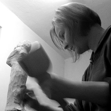 Photographie en noir et blanc de l'artiste et sculptrice allemande Angelika Kienberger en train de travailler à la main, à l'aide d'un marteau et d'un ciseau, sur une sculpture en bois.
