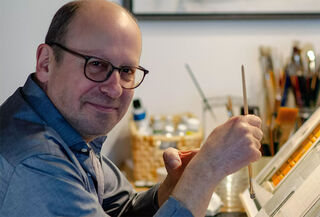 Der Maler Ralf Kunstmann blickt lächelnd mit Brille von seinem Zeichentisch auf und hält einen feinen Pinsel.