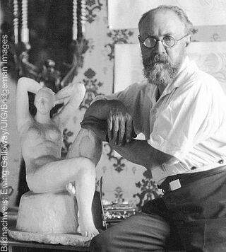 Historische Schwarz-Weiß-Fotografie des Malers Henri Matisse mit Brille und Bart neben einer Frauenskulptur.