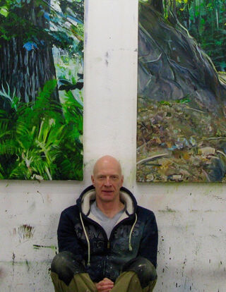 L'artiste et peintre allemand Burkhard Kern, chauve, assis dans son atelier devant deux de ses peintures naturalistes grand format représentant des forêts et des paysages.