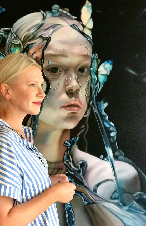 Umělkyně Valentina Andrees stojí v popředí v profilu a dívá se na jeden ze svých velkoformátových surrealistických obrazů, který zachycuje ženskou tvář s modrými motýly.