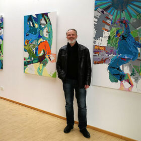 L'artiste Uwe Fehrmann se tient dans une galerie devant deux tableaux colorés.