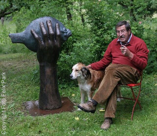 Lo scrittore, artista, pittore e scultore tedesco Günter Grass, seduto in giardino con una pipa in mano su una sedia accanto a un cane, mentre tiene in mano la sua scultura “Butt”.