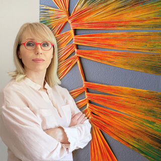 Alla Grande is standing with her arms crossed in front of an orange-and-yellow relief.