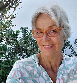 A close-up of the artist and painter Mo Kilders, wearing glasses and with grey hair, smiling warmly outdoors in front of some green plants.