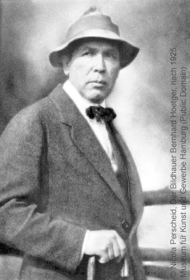 Portrait historique en noir et blanc de l'artiste et sculpteur allemand Bernhard Hoetger, coiffé d'un chapeau, vêtu d'un costume et d'un nœud papillon, s'appuyant sur une canne.