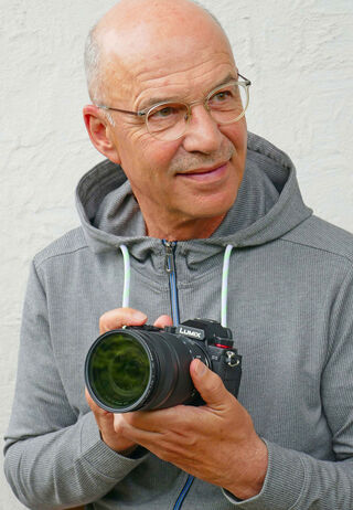 Portrait du photographe Gottfried Römer, portant des lunettes, qui tient un appareil photo Lumix entre ses mains.